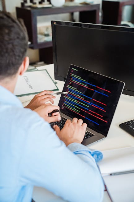 A person in a light blue shirt sitting at a desk during a home relocation process, working on a silver laptop with a screen displaying colorful lines of computer code. The workspace includes a closed