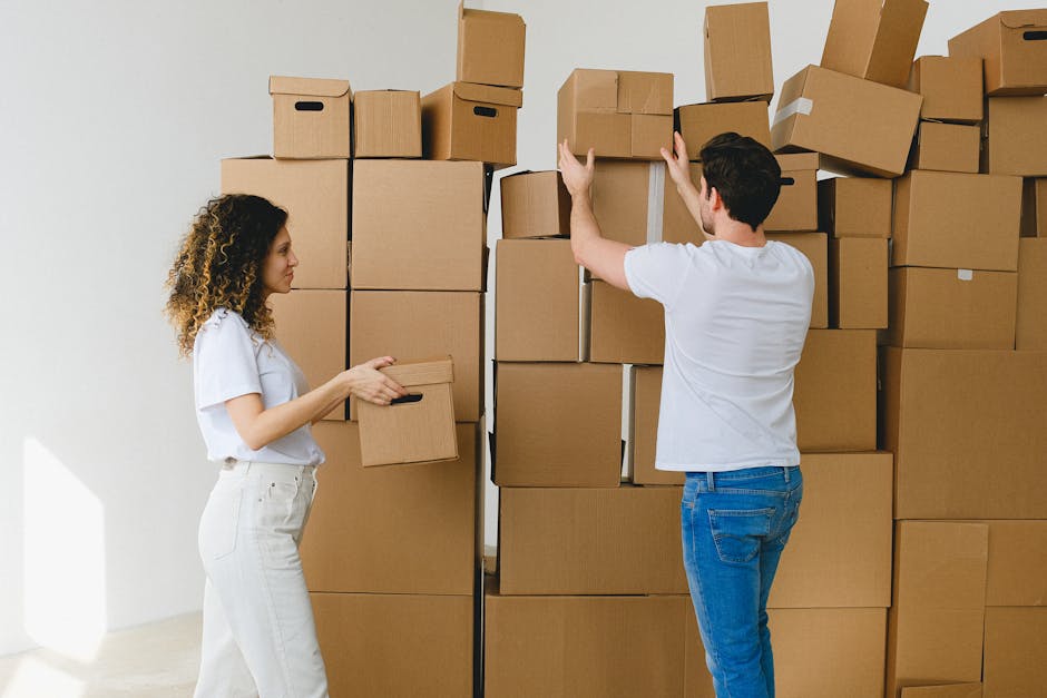 A man and a woman are inside a room surrounded by tall stacks of cardboard boxes, some open and others sealed with white packing tape. The woman, with curly hair, dressed in a white t-shirt and white 