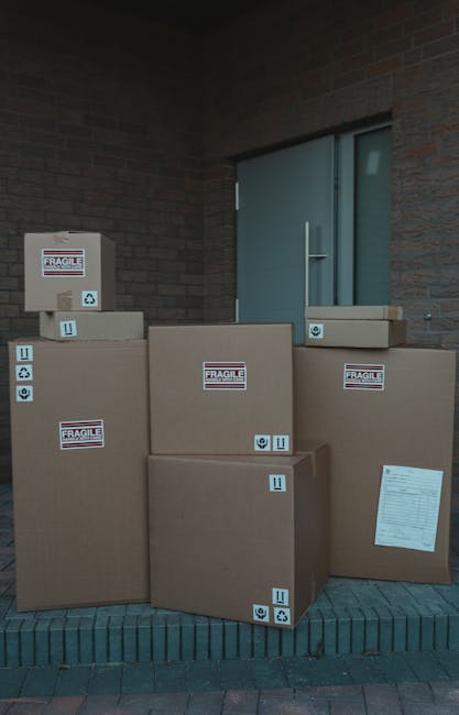 A man and a woman are inside a room surrounded by tall stacks of cardboard boxes, some open and others sealed with white packing tape. The woman, with curly hair, dressed in a white t-shirt and white 